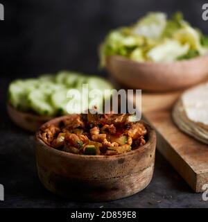 Eine Schüssel mit einem Huhn Fleisch und Gemüse Füllung auf einem Tisch, neben einem Teller mit einigen Scheiben Gurke, eine Schüssel mit Salat und einige Tortillas oder Stockfoto