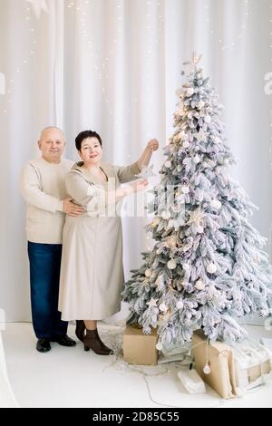Mittleres Alter glückliches Paar, das sich auf die Winterferien vorbereitet, schönen Weihnachtsbaum im gemütlichen Lichtstudio oder zu Hause dekoriert. Neujahr Stockfoto