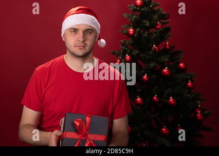 Junger Mann in Weihnachtsmann Hut rot T-Shirt hält Geschenk Box mit Weihnachtsgeschenk Stockfoto