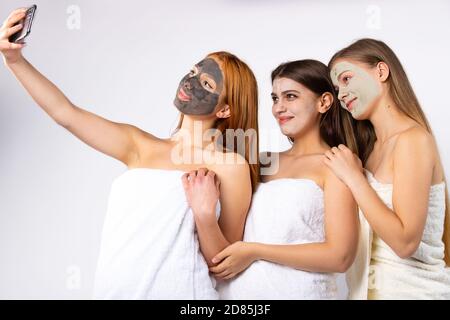 Drei junge glückliche Mädchen mit Gesichtsbehandlung, eingewickelt in Handtücher, nehmen Sie ein Selfie. Beauty- und Entspannungskonzept. Foto auf weißem Hintergrund Stockfoto