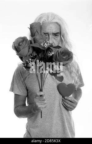 Studioaufnahme eines älteren bärtigen Mannes, der sich hinter Rosen versteckt Halten Sie das Herz bereit für den Valentinstag Stockfoto