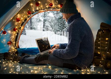 Mann sitzt im Zelt mit Weihnachtsbeleuchtung geschmückt, mit Laptop. Schöne Draufsicht auf skandinavische Landschaft, mit Schnee bedeckter Wald. Stockfoto