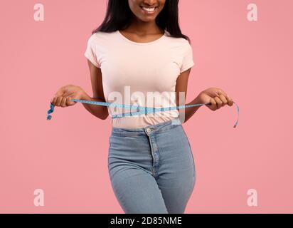 Schlankheits- und Diät-Konzept. Unkenntlich schwarze Frau misst ihre Taille während der Gewichtsabnahme über rosa Hintergrund Stockfoto