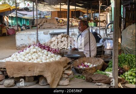 Nagpur, Maharashtra, Indien - März 2019: Ein indischer Gemüseverkäufer mit Haufen Knoblauch, Zwiebeln und Kartoffeln zum Verkauf in seinem Laden in einer Gemüsemarke Stockfoto