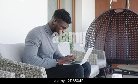Der junge schwarze Mann, der auf seinem Balkon am Laptop arbeitet, während er Kaffee trinkt. Hochwertige Fotos Stockfoto