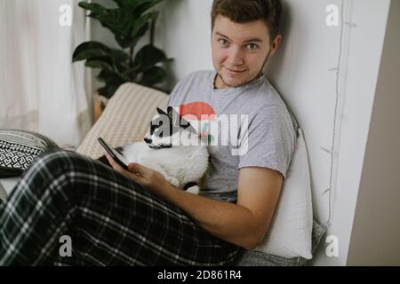 Schöner Mann, der mit einer Katze sitzt und das Telefon in der Hand hält, in einem stilvollen, modernen Zimmer. Nette Katze spielt mit Besitzer und zieht die Aufmerksamkeit von Smartphone Stockfoto