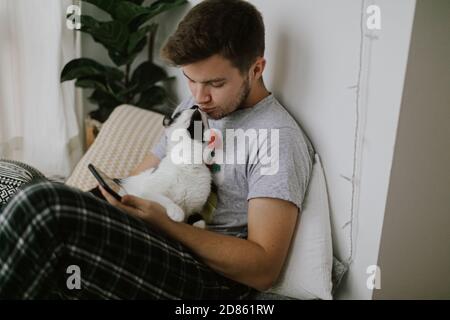 Schöner Mann, der mit einer Katze sitzt und das Telefon in der Hand hält, in einem stilvollen, modernen Zimmer. Nette Katze spielt mit Besitzer und zieht die Aufmerksamkeit von Smartphone Stockfoto