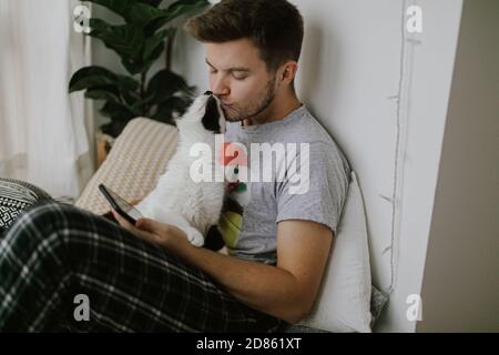 Nette Katze spielen und küssen Besitzer und die Aufmerksamkeit von Smartphone. Schöner Mann mit Katze sitzen und halten Telefon in der Hand in stilvollen modernen Stockfoto