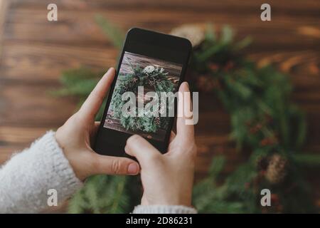 Hände halten Telefon und Foto von stilvollen rustikalen weihnachtskranz auf Holztisch, saisonalen Winter Online-Workshop. Handgemachte festliche Einrichtung, Holi Stockfoto