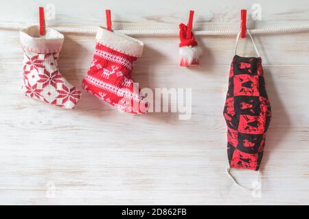 Sicheres Weihnachtskonzept. Weihnachtssocken, kleiner Weihnachtsmann auf Fakemask über weißer Holzwand. Weihnachtsstrumpf, Weihnachtsmann Spielzeug auf schützende Gesichtsmaske h Stockfoto