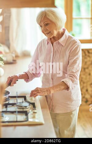 Lächelnde ältere Frau, die den Herd anmacht und die Bratpfanne anlegt, um zu Hause zu frühstücken. Inländisches Lifestyle-Konzept Stockfoto