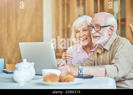 Glücklicher älterer Mann und Frau umarmen, während sie am Tisch sitzen und auf den Laptop-Bildschirm schauen. Inländisches Lifestyle-Konzept Stockfoto
