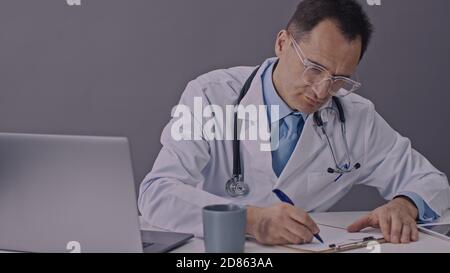 Fokussierter Arzt arbeitet mit Zwischenablage schreibt Krankenakte müde legt Am Schreibtisch Stockfoto