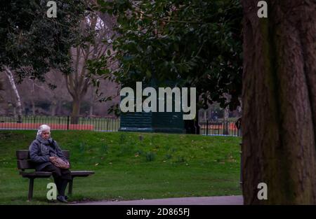 London, Vereinigtes Königreich - APR 12, 2019 : Ältere kaukasische Frau sitzt auf Holzstuhl in Parkbank. Kein Fokus, speziell. Stockfoto