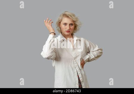 Junge blonde Frau in einem weißen Hemd sieht die Kamera an, Hand an Hand, andere Hand angehoben. Überraschte Pose. Stockfoto