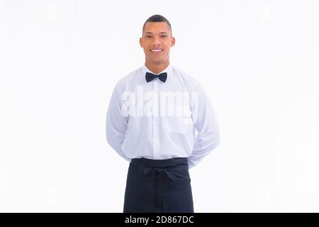 Hübscher junger Kellner, der mit den Händen hinter dem Rücken auf einem hellen Hintergrund aufsteht. Arbeits- und Servicekonzept. Stockfoto
