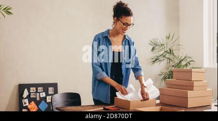 Frau, die den Karton zu Hause auf ihren Schreibtisch packt. Geschäftsfrau, die das Produkt für die Lieferung an den Kunden vorbereitet. Stockfoto