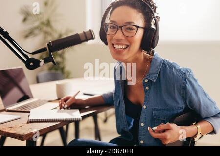 Fröhliche weibliche Podcast-Host arbeitet von zu Hause aus. Frau trägt Kopfhörer sitzen am Tisch mit Laptop, Mikrofon und einem Notizblock Blick auf Kamera und Stockfoto