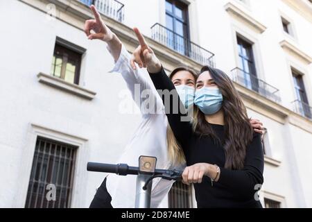 Zwei lustige Mädchen zeigen ihre Hände in eine Richtung. Sie stehen auf einem Elektroroller und tragen Gesichtsmasken. Stockfoto