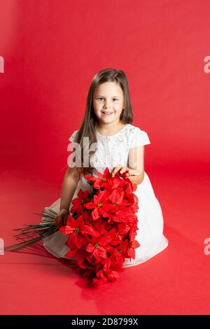 Kind in weißem Kleid sitzt auf dem Boden und hält einen Strauß Weihnachtsblumen auf ihrem Schoß, isoliert auf einem roten Hintergrund Stockfoto