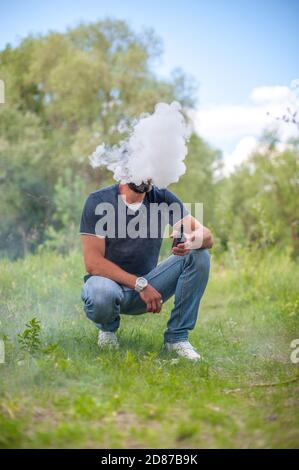 Bärtiger Mann bläst viel Rauch mit Vape elektronische Zigarette auf dem Waldboden. Verdampfungsaktivität. Stockfoto