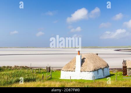 Traditionell gebaute weiße Hütte mit Strohdach, neben der türkisfarbenen Bucht, mit stürmischen bewölkten dunklen Himmel über.Insel North Uist, Schottland Stockfoto