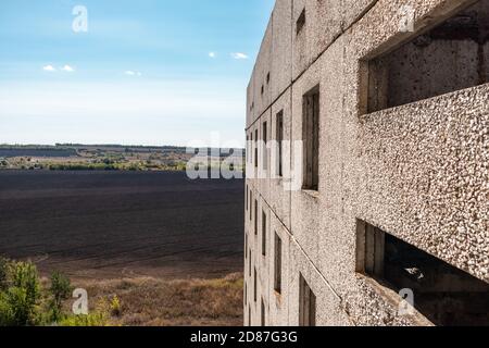 Blick aus dem Fenster des hohen mehrstöckigen verlassenen sowjetischen Gebäudes mit Fassade auf hellblaue ländliche Landschaft. Verwitterte Wohnsiedlung, unvollendete Stadt o Stockfoto