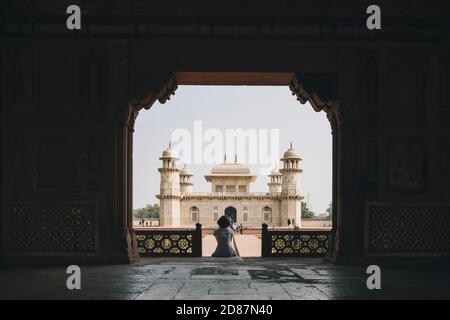 Junge Frau am Eingangstor des Grabes von Itmad-ud-Daula mit dem Mausoleum im Hintergrund sitzend. Stockfoto