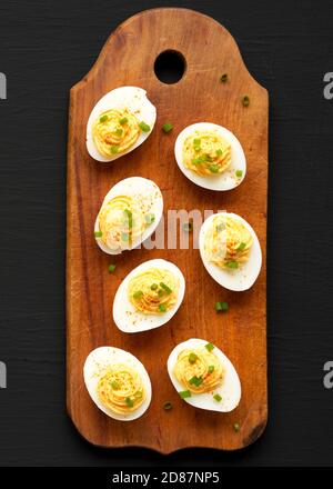 Hausgemachte Deviled Eier mit Schnittlauch auf einem rustikalen Holzbrett auf einer schwarzen Oberfläche, Draufsicht. Flachlage, über Kopf, von oben. Stockfoto