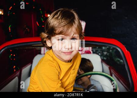 Netter kleiner Junge, der auf rotem Weihnachtsauto sitzt und die Kamera anschaut. Kind. Weihnachtsstimmung. Feiertage. Weihnachten. Neujahr. Stockfoto