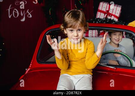 Netter kleiner Junge, der auf rotem Weihnachtsauto sitzt und die Kamera anschaut. Kind. Weihnachtsstimmung. Feiertage. Weihnachten. Neujahr. Stockfoto