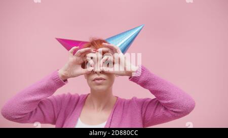 Verrückte Frau mittleren Alters mit 2 Party Hüte und Finger bilden Brille über ihre Augen isoliert auf rosa Hintergrund. Hochwertige Fotos Stockfoto