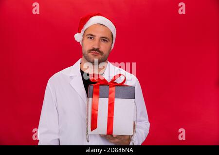 Müde Arzt in Weihnachtsmann Hut hält Weihnachtsgeschenk-Box Blick auf die Kamera Stockfoto