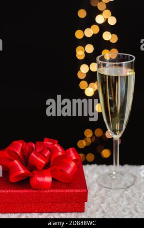 Rote weihnachts-Geschenkbox mit verschwommenem Fokus auf einem Glas Sekt und goldenen Bokeh-Lichtern auf dunklem Hintergrund. Stockfoto