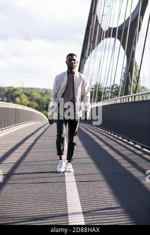 Junger Mann, der mit Zuversicht auf der Fußgängerbrücke läuft. Volle Länge. Vorderansicht. Lange Aufnahme. Stockfoto
