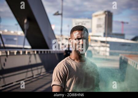 Junger schwarzer Mann, der die Kamera anschaut und grünen Rauch aufsteigt. Mittlere Aufnahme. Stockfoto