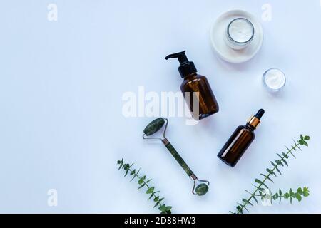 Gesichtswaschgel und Serum in dunklen Glasflaschen, Cremeglas, Gesichtswalze, Eukalyptuszweige auf hellblauem Hintergrund. Natürliche Bio-Kosmetik Konz Stockfoto