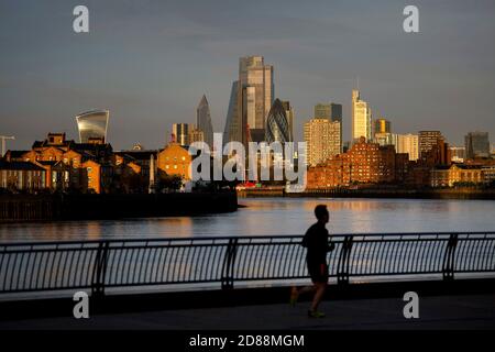 Ein Jogger am frühen Morgen im Osten Londons läuft an der Skyline der City of London vorbei, die von Sonnenaufgang beleuchtet wird. Stockfoto