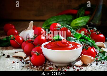 Tomatenketchup-Sauce mit Knoblauch, Gewürzen und Kräutern mit Kirschtomaten in einer weißen Porzellanschüssel auf altem Holztisch, selektiver Fokus Stockfoto