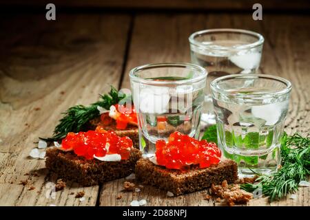 Kalter russischer Wodka mit Eis und kleinen Snacks Sandwiches mit Butter und rotem Lachskaviar auf einem alten Holztisch im rustikalen Stil, selektiver Fokus Stockfoto