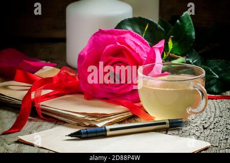 Vintage Postkarte mit Tasse Kräutertee, ein Päckchen alte Buchstaben, gebunden mit einem Band, rosa Rose, Kerzen auf dem alten Holzhintergrund, selektiv Stockfoto