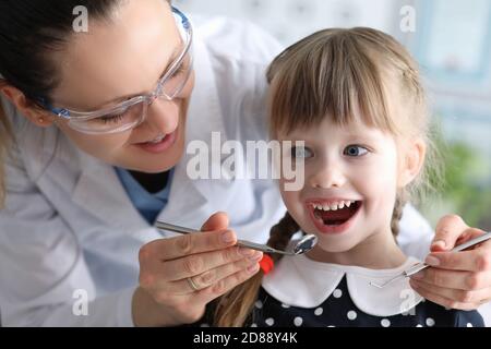 Frau Zahnarzt in Schutzbrille untersucht Zähne des kleinen Mädchens Patient mit Metallwerkzeugen im Hochformat Stockfoto