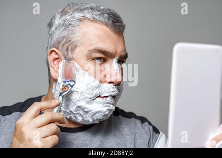 Grauhaariger Mann schaut in den Spiegel und rasiert Stockfoto