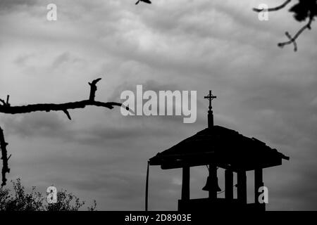 Selektiver Fokus auf metallischen Kreuz auf kleine alte Kirche Glockenturm am entfernten orthodoxen Tempel, Südbulgarien, bewölkten Herbsttag. Moody Darkness Silhouetten Bild Stockfoto