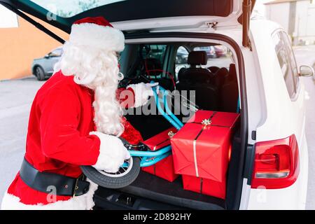 Porträt des Weihnachtsmannes mit dem Kofferraum des Autos Öffnen Sie voll von Geschenken, die einen Kinderroller mit Große Räder, um die Geschenke zu liefern, weil seine Stockfoto