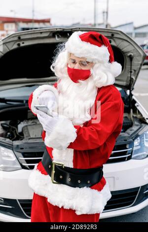 Porträt des Weihnachtsmannes, der etwas in sein Handy schreibt Besorgt und mit der Haube seines Autos geöffnet, weil Sein Auto ist kaputt gegangen und er kann nicht d Stockfoto