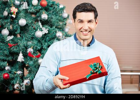 Lächelnder Mann mit einem roten Weihnachtsgeschenk neben einem Weihnachtsbaum Stockfoto