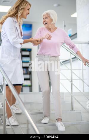 Doktor in einem Laborkittel, der ihre Patientin bei der unterstützt Treppen und sieht freundlich aus Stockfoto