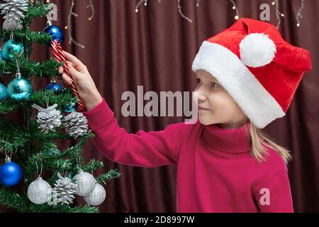 Ein süßes kaukasisches blondes Mädchen in einem roten Pullover und einer Weihnachtsmütze hängt Weihnachtsbonbons auf einem geschmückten Weihnachtsbaum. Weihnachts- und Neujahrskonzept Stockfoto