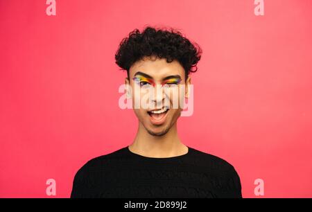 Gay Mann mit Regenbogen Auge Make-up winken ein Auge vor rotem Hintergrund. Geschlecht Flüssigkeit männlich winken bei der Kamera. Stockfoto